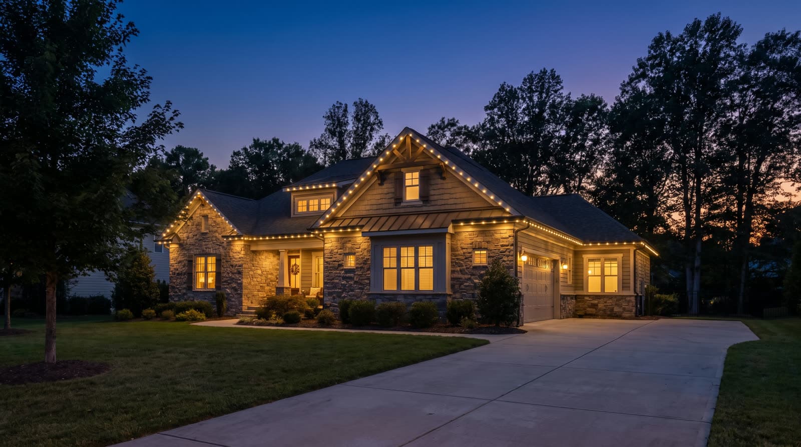 Stone and shingle craftsman house at twilight with warm white roofline lighting tracing every gable, tall trees framing the lot