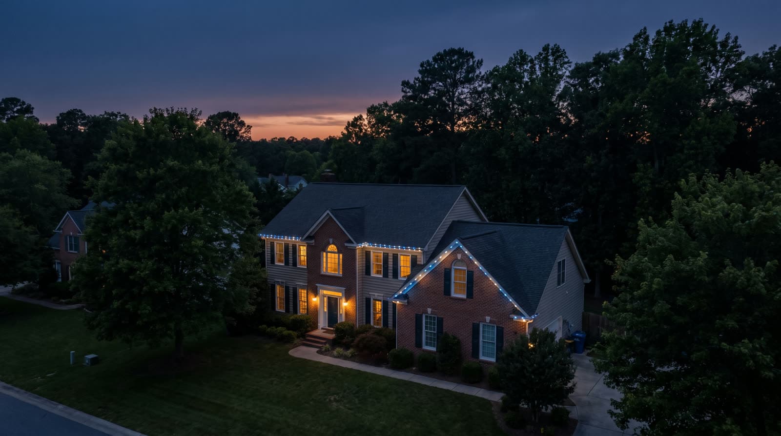 Aerial view of a two-story brick-and-tan-siding colonial tucked among tall trees after sunset, cool white lights tracing both roof gables and the main eave, lit windows on both floors, a neighboring rooftop visible at left, a dark tree line and fading orange horizon band in the background.