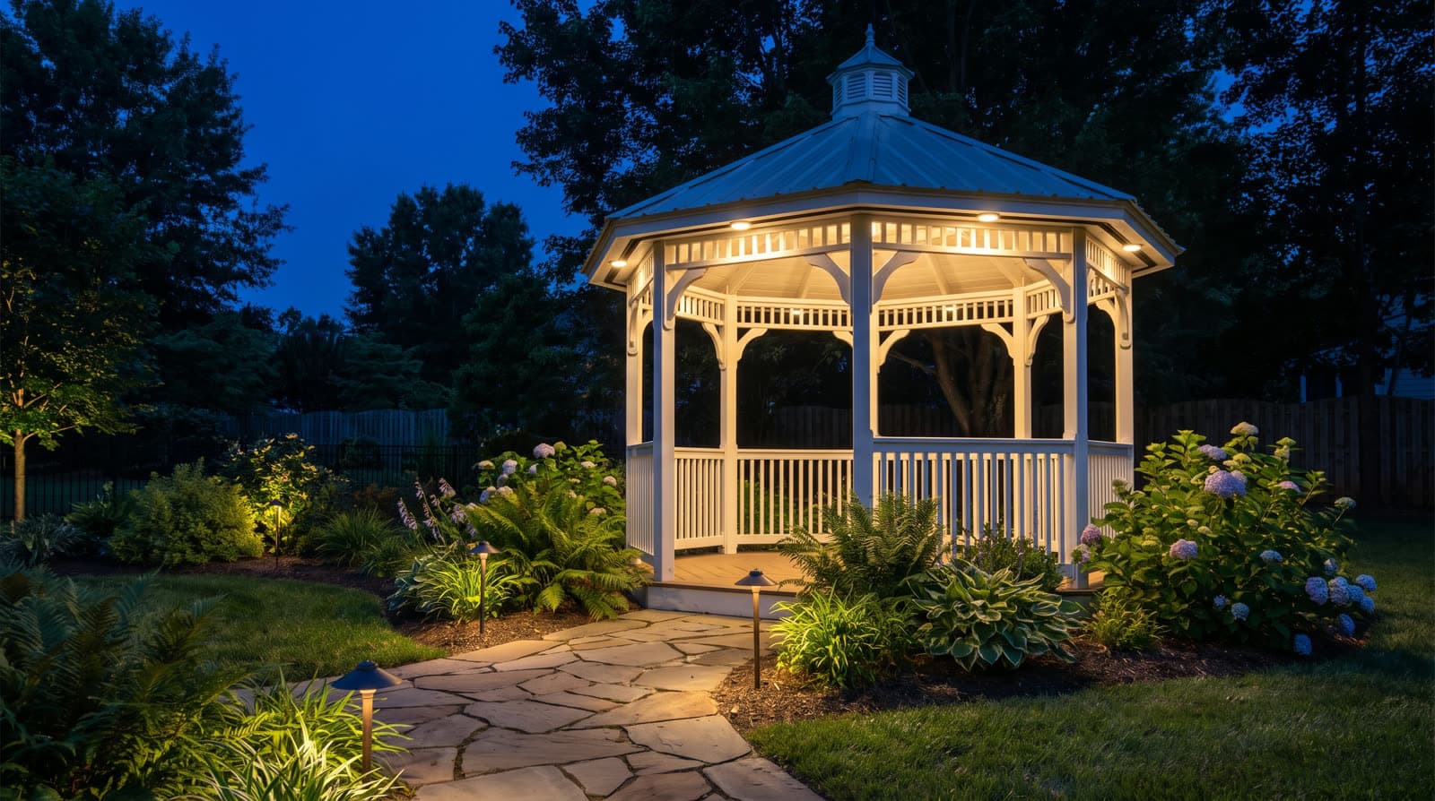 White octagonal gazebo at night with warm downlights under its roof, flagstone path lit by low path lights, hydrangeas in the beds around it