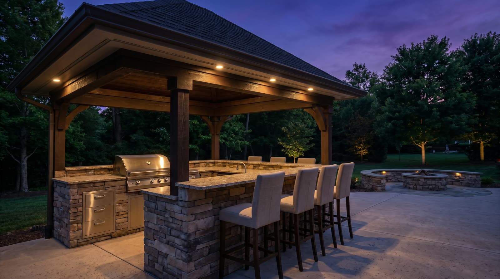 Covered outdoor kitchen with stone bar, grill and bar stools at twilight, round downlights in the pavilion ceiling, a fire pit and illuminated seat wall beyond