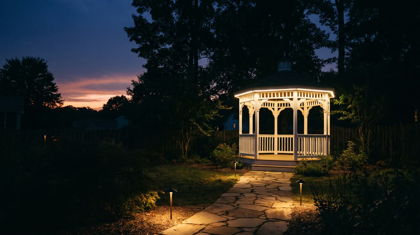 White gazebo glowing at night at the end of a flagstone path, warm outline along its roof edge, path lights leading to it through the trees