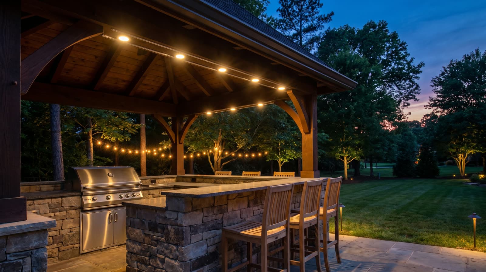 Covered outdoor kitchen with stone bar and grill at twilight, round downlights in the wood ceiling, a strand of warm bistro lights beyond, path lights on the lawn