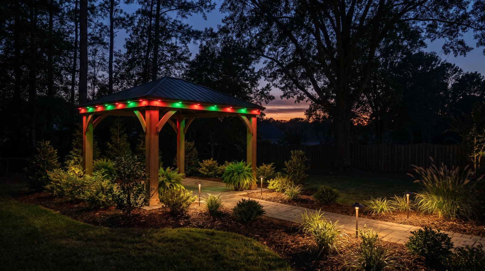 Small wood pavilion in a garden at twilight, red and green lights along its roof edge, path lights along a walkway through planted beds