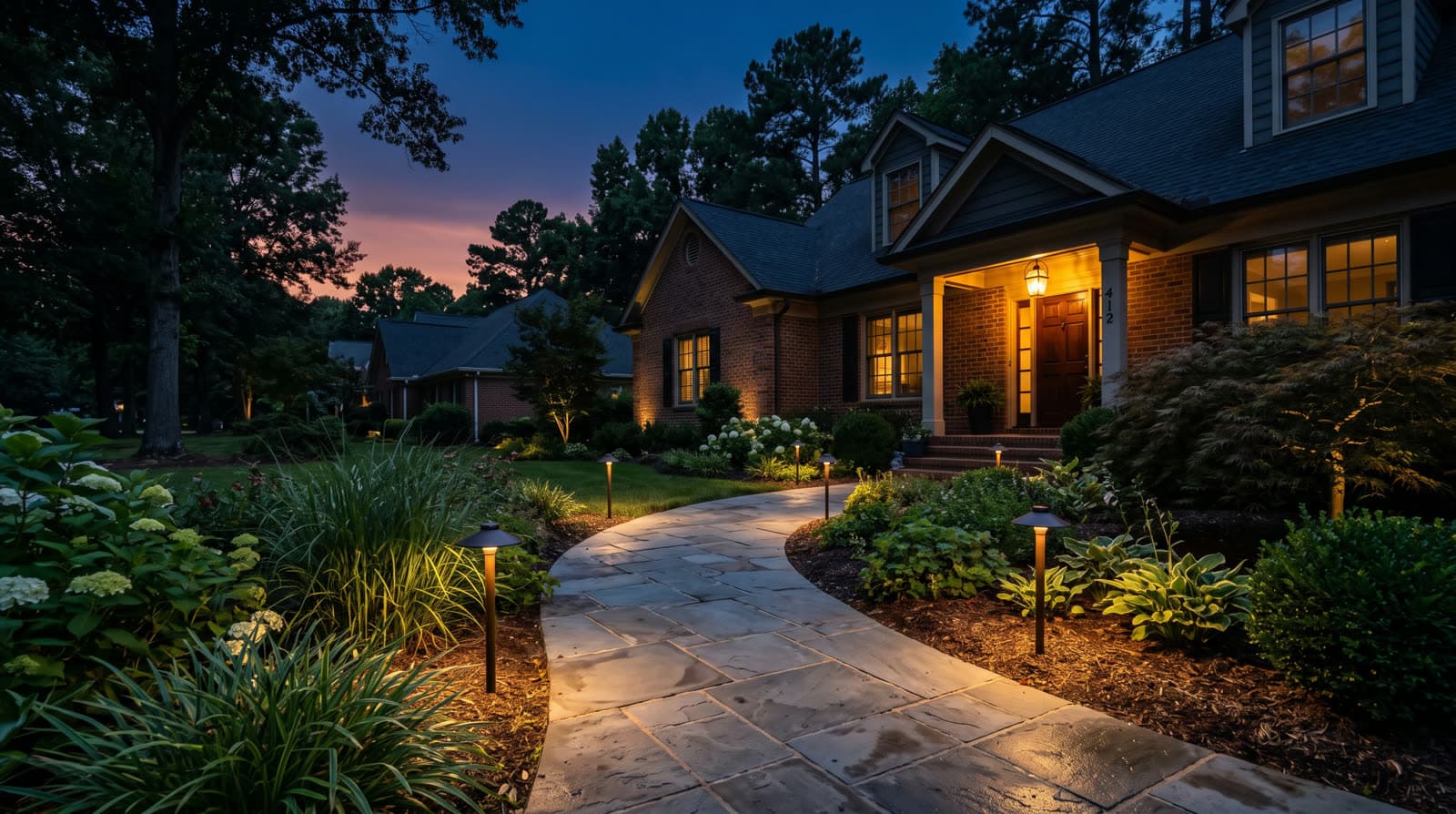 Brick house with lit front porch at twilight, curving stone walkway bordered by hostas and hydrangeas, dark bronze path lights spaced along both sides