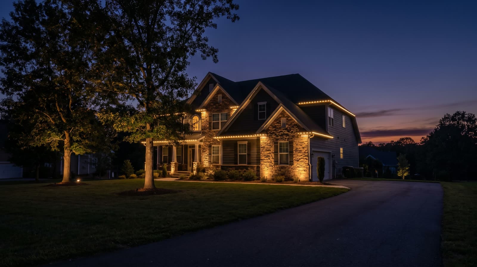 Two-story house with dark siding, stacked-stone accents and a covered front porch on square columns, photographed from across a curved driveway after sunset, warm white lights running the full length of the lower and upper eaves and the porch roof line, lit windows on both floors, two large shade trees at left, a deep blue sky fading to orange at the horizon.