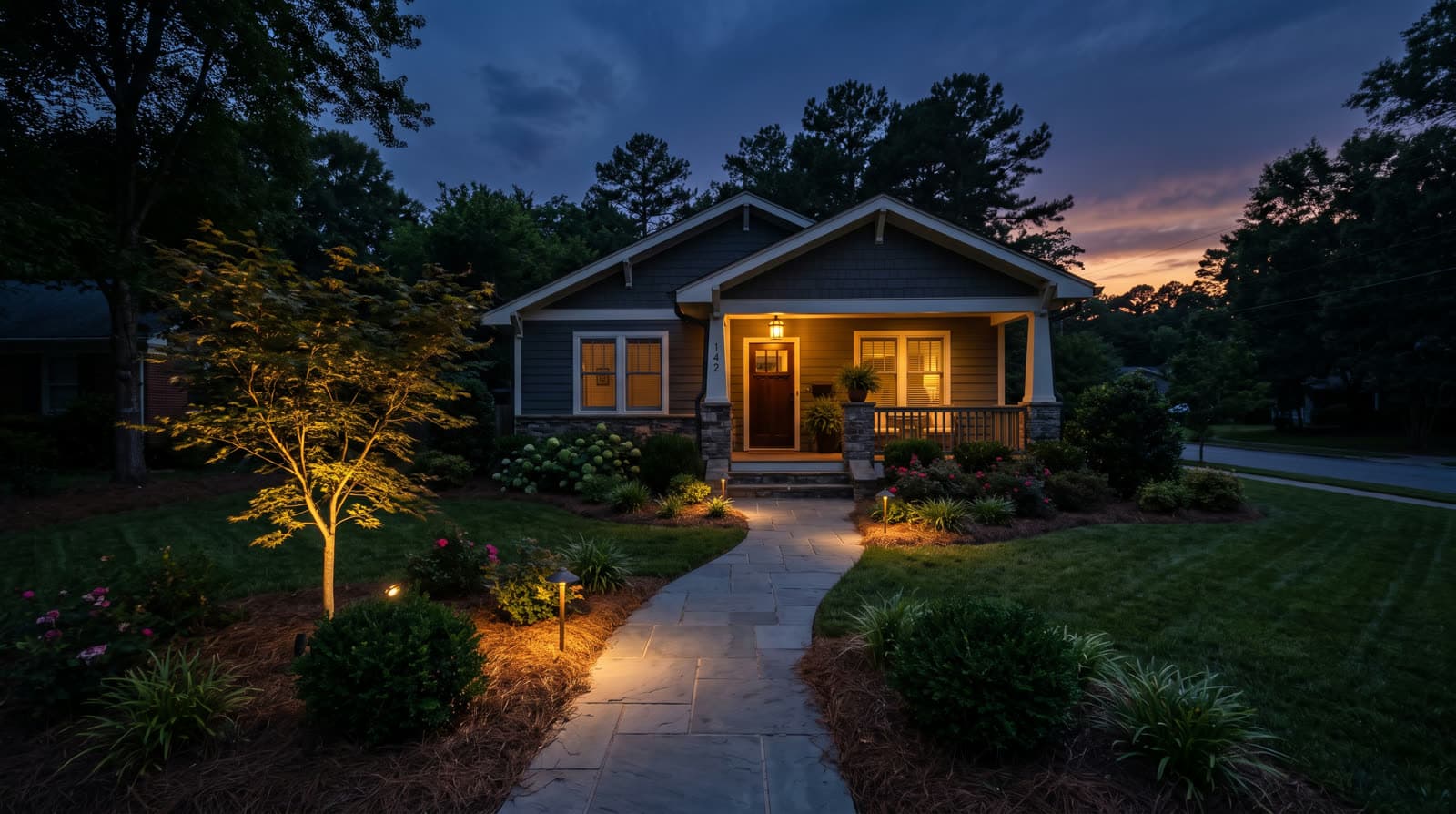 Craftsman bungalow with covered front porch at twilight, stone walkway lined with low path lights to the steps, small tree uplit in the front bed